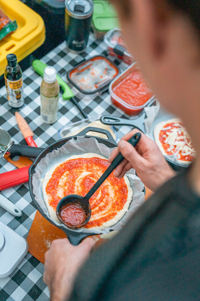 Man is spreading pizza sauce on pizza in a cast iron at camp.